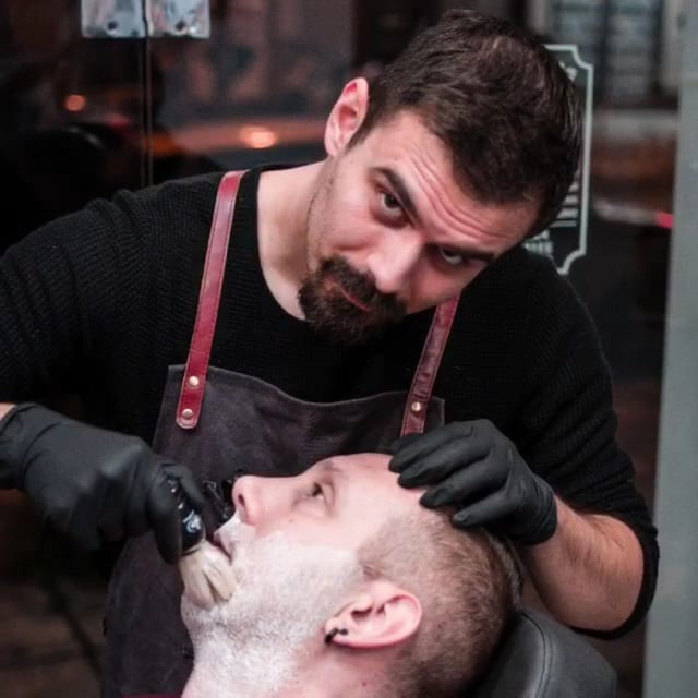 Ο κουρέας ξυρίζει πελάτη με φαλτσέτα, με μαύρα γάντια και δερμάτινη ποδιά